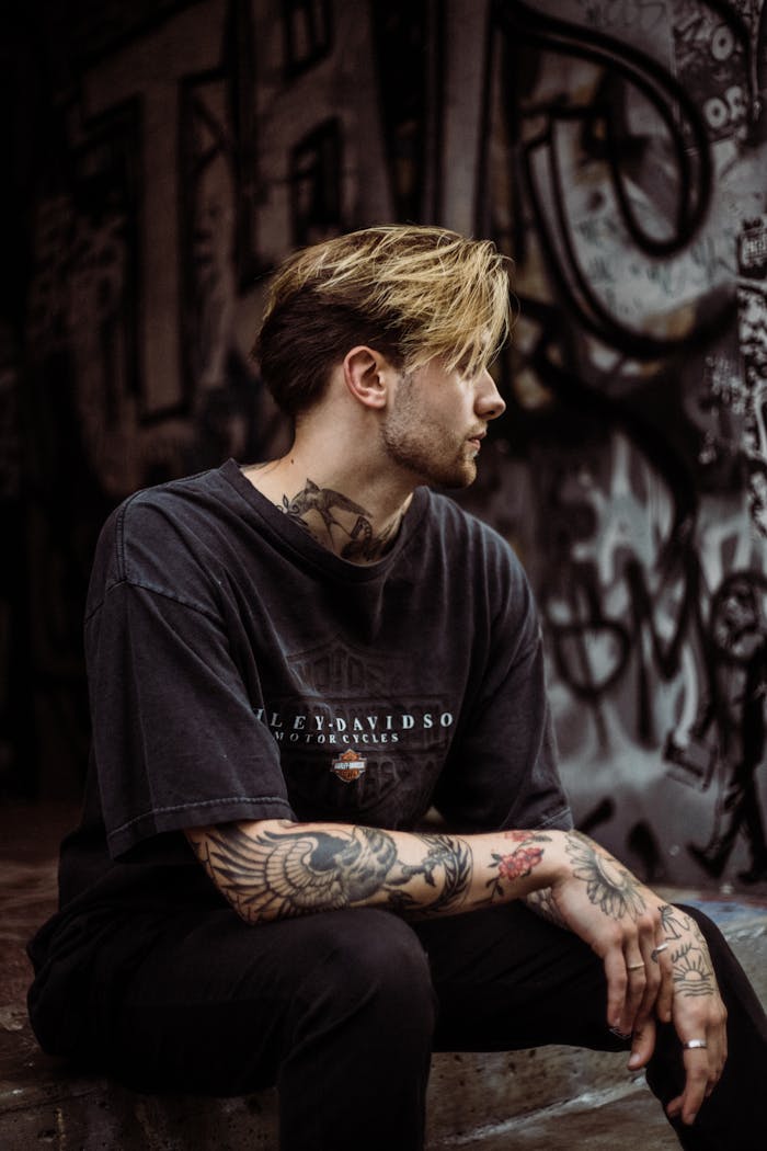 about-img-02 A tattooed man in a black T-shirt sits in a graffiti-covered urban environment, showcasing street style.